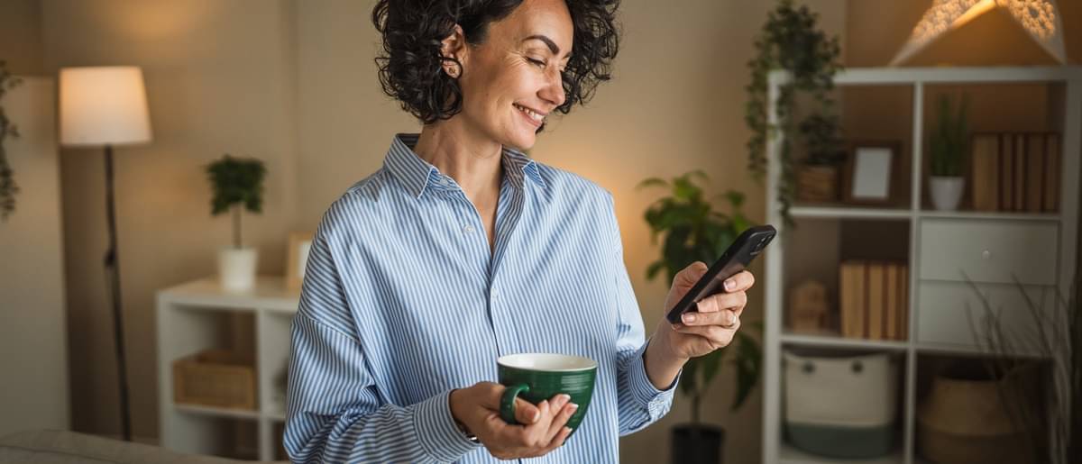 Eine Frau steht lässig mit einer Tasse in der Hand im Raum und schaut auf ihr Smartphone