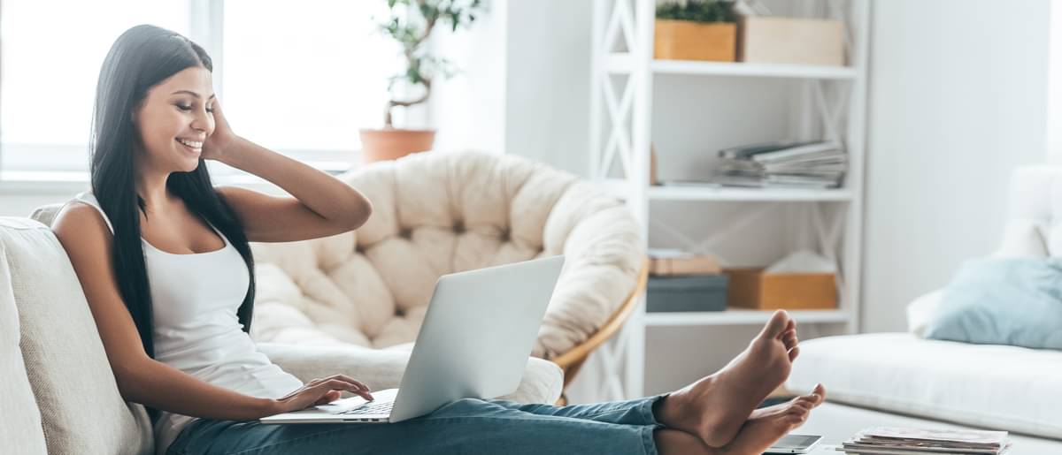 Eine lächelnde junge Studentin sitzt mit ihrem Laptop auf dem Sofa und hat ihre Füße auf dem Couchtisch hochgelegt. - Studium finanzieren