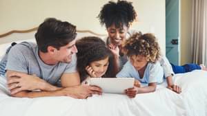 Eltern liegen gemütlich mit ihren zwei Kindern auf dem Bett und schauen auf ein Tablet.