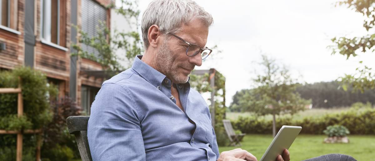 Mann mit grauen Haaren und Brille in Hemd sitzt entspannt auf Gartenstuhl vor Haus und tippt auf Tablet.