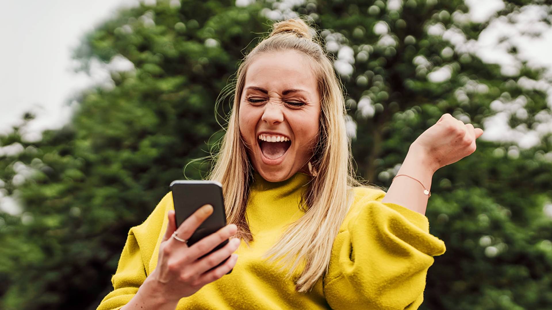 Eine junge, blonde Frau mit Zopf und gelben Pulli schaut auf ihr Handy und freut sich und hebt dabei einen Arm mit der Hand zu Faust geballt nach oben.
