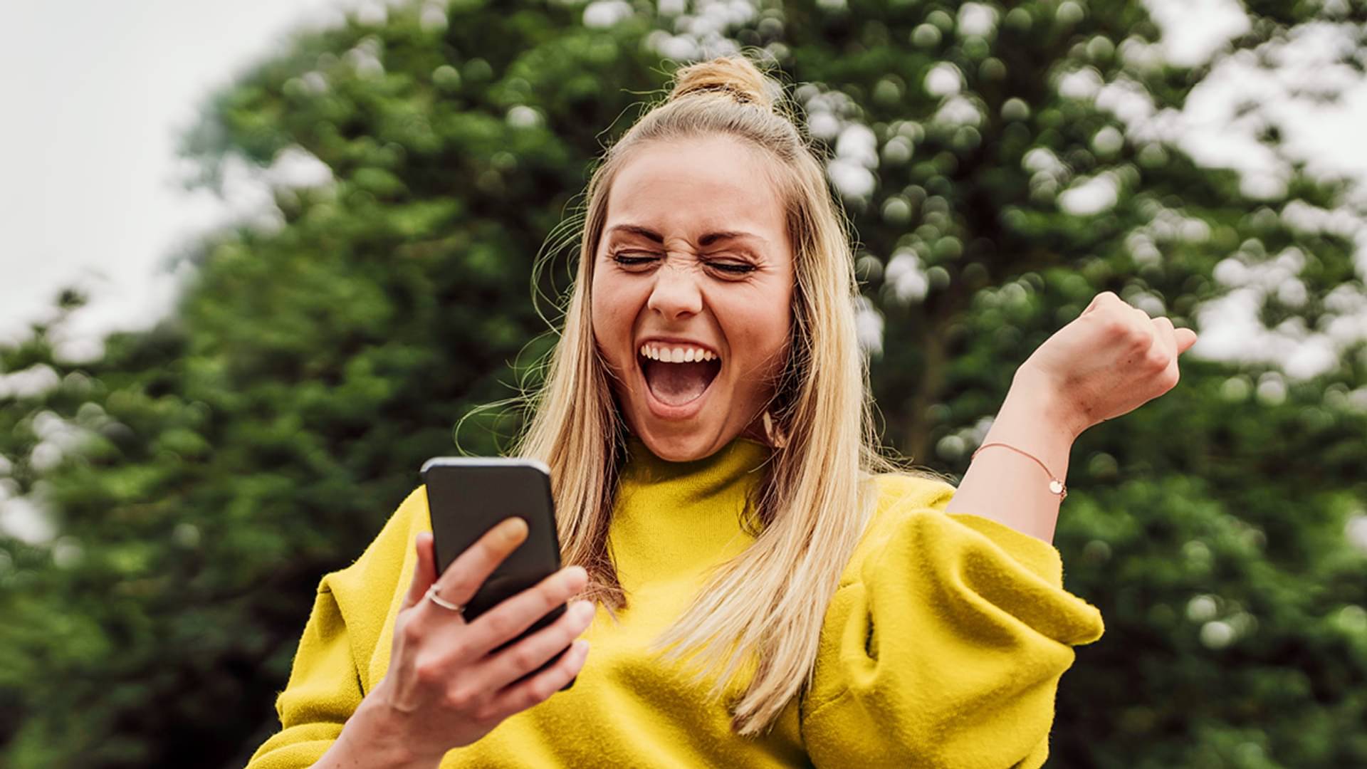 Eine junge, blonde Frau mit Zopf und gelben Pulli schaut auf ihr Handy und freut sich und hebt dabei einen Arm mit der Hand zu Faust geballt nach oben.