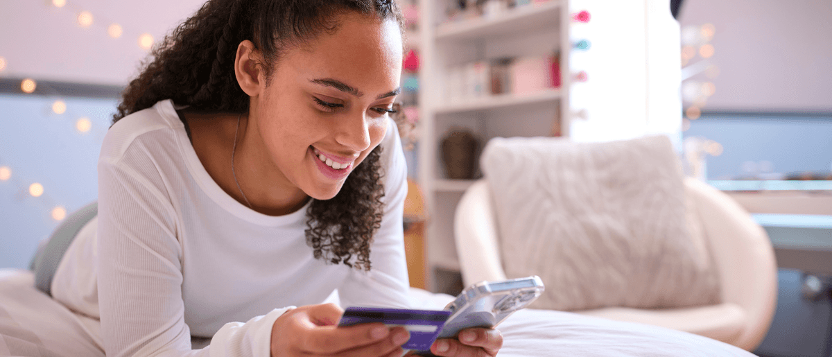 Eine fröhliche Teenagerin liegt auf dem Bett und schaut auf ihr Handy. In der anderen Hand hält sie eine Bankkarte.