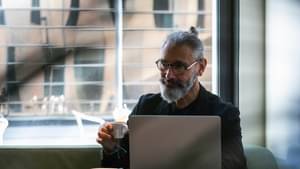Älterer, eleganter Mann sitzt in einem Café und checkt sein Premiumdepot an einem Laptop.