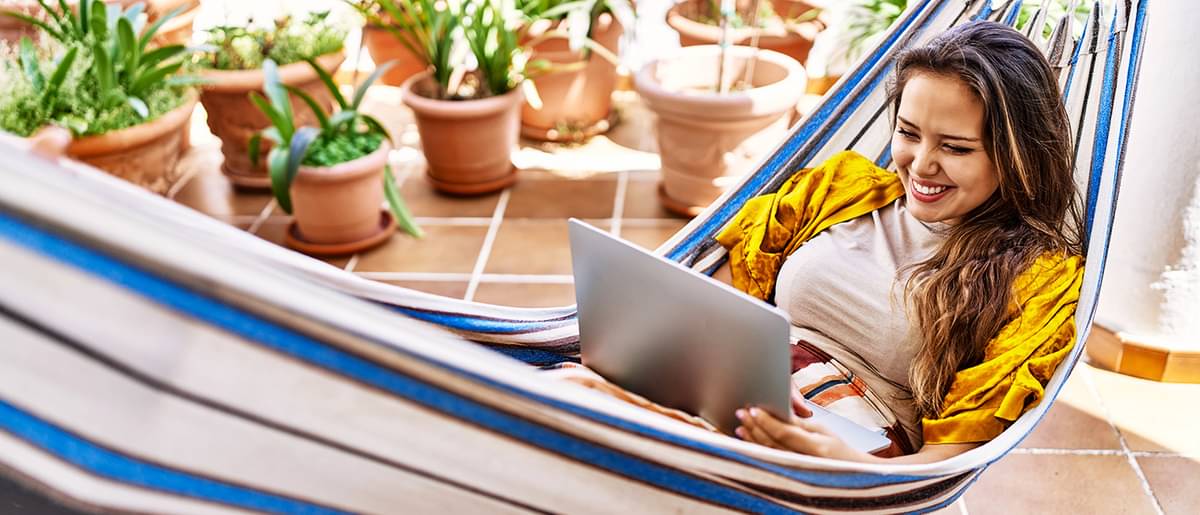 Eine junge Frau sitzt fröhlich mit dem Laptop auf dem Schoß in einer Hängematte auf der Terrasse und informiert sich über Kreditarten.